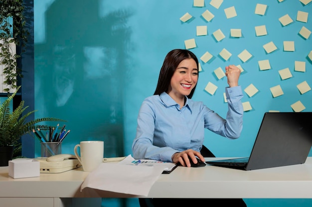 Оптимистичная азиатка, сидящая за офисным столом, рада получать хорошие новости о проекте по электронной почте. улыбающийся корпоративный сотрудник празднует результат ежемесячной статистики на рабочем месте.