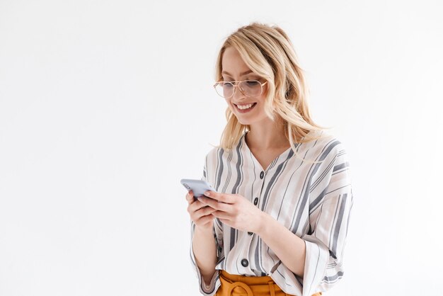 Donna attraente e ottimista che indossa occhiali che sorride al telefono e scrive isolata sul muro bianco