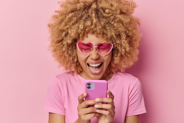 Optimistic curly haired woman screams from joy keeps eyes closed watches something funny on smartphone glad to buy new cellphone dressed in casual t shirt heart shaped pink shades poses indoor