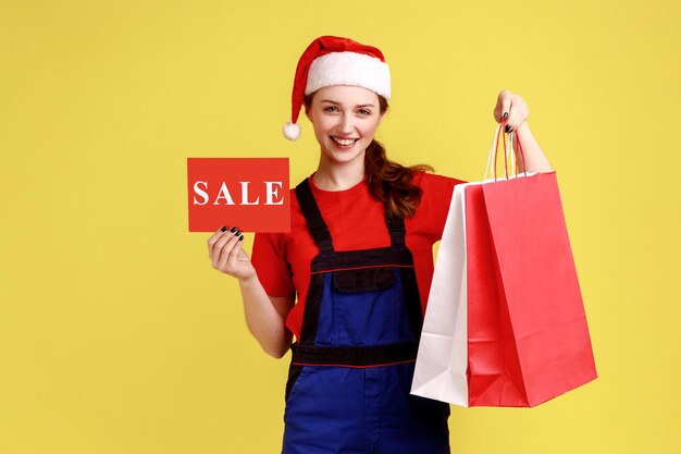 Optimistic delivery woman shows card with sale inscription and shopping bags discounts for shipping wearing blue overalls and santa claus hat Indoor studio shot isolated on yellow background