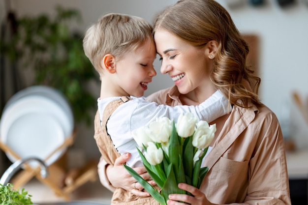Фото Оптимистичная семейная мать с букетом тюльпанов улыбается и касается лба счастливому сыну во время празднования дня матери дома