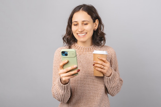楽観的な女性は、電話でブラウジングまたはテキストメッセージを送信しながらテイクアウトコーヒーを楽しんでいます