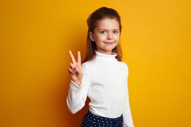 Optimistic girl kid showing peace gesture against bright yellow background