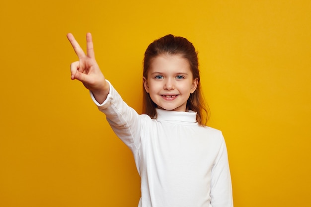 Оптимистичный ребенок-девочка показывает жест мира на ярко-желтом фоне