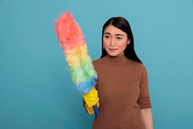 Optimistic glad asian housewife in yellow rubber gloves holding dust brush isolated on a blue background, Cleaning home concept, Feeling Cheerful happy and positive