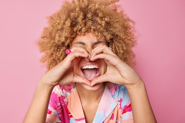 Optimistic glad woman with curly bushy hair makes heart gesture over mouth says I love you shows romantic gesture dressed in colored shirt isolated over pink background Happy Valentines Day