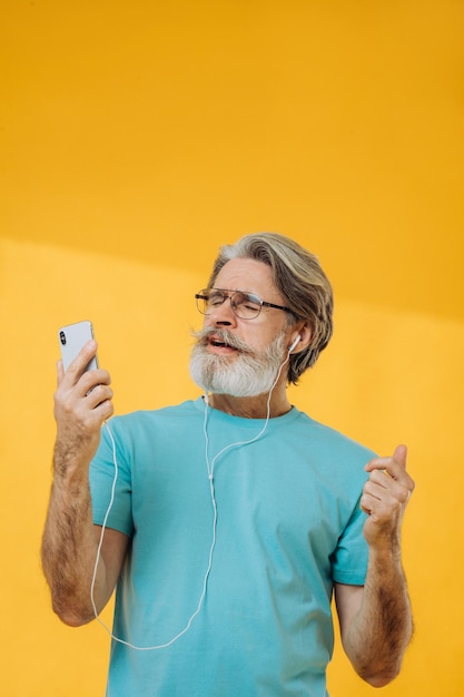 낙천적인 회색 머리 노인은 노란색 배경에 격리된 헤드폰 안경을 쓰고 음악 춤을 듣습니다.
