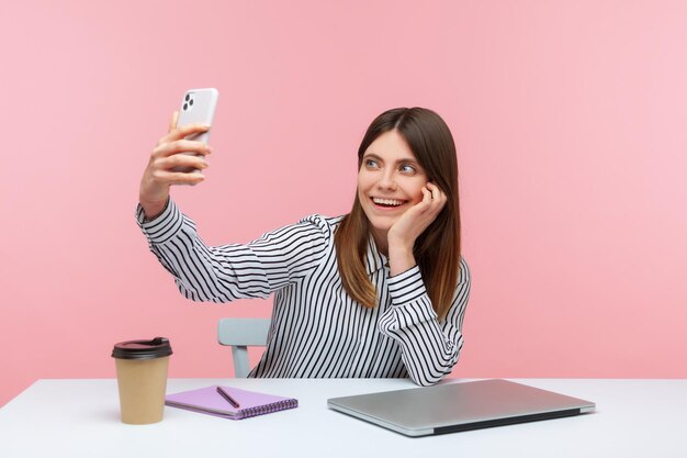 職場に座っているスマートフォンのカメラで自分の写真を撮って、楽しい気分で笑っている楽観的なポジティブな女性サラリーマンピンクの背景に分離された屋内スタジオショット