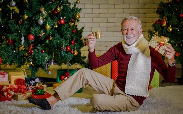 Optimistic senior man with knitted scarf looking at camera with smile and showing presents while sitting on carpet near decorated Christmas trees against brick wall