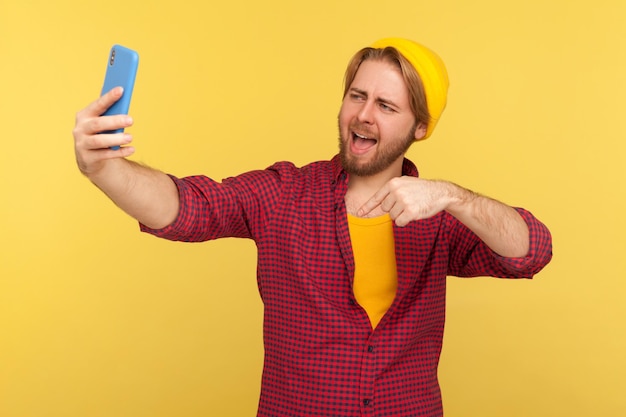 Optimistic trendy hipster guy in checkered shirt pointing\
finger down click below smiling talking video call or streaming on\
mobile phone asking follow and subscribe his blog studio shot\
isolated