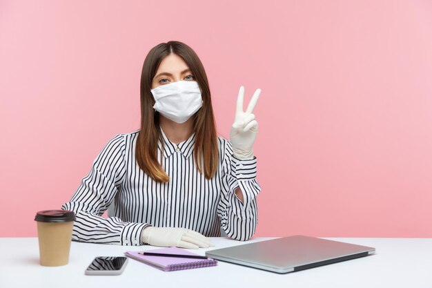 Photo optimistic woman employee sitting safe healthy with protective mask and gloves during quarantine, showing victory, peace gesture. working at home office in self-isolation, coronavirus outbreak. indoor