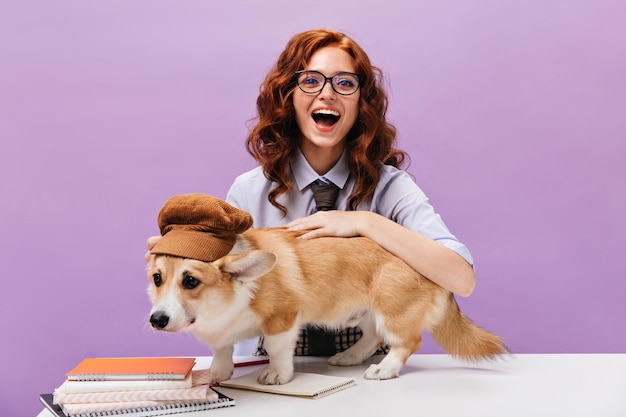 Optimistic woman in eyeglasses smiles and hugs corgi Funny student girl in shirt plays with dog in cap on isolated backdrop