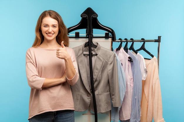 Optimistic woman, fashion stylist standing near business clothes hang on shelf in designer store and showing thumbs up, selling trendy outfit collection in shop boutique. indoor studio shot isolated