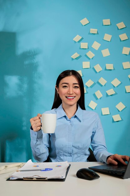 Optimistic woman sitting at workstation drinking a cup of coffee while working with a laptop. Casual startup employee relaxing at work with statistics in a business workplace.