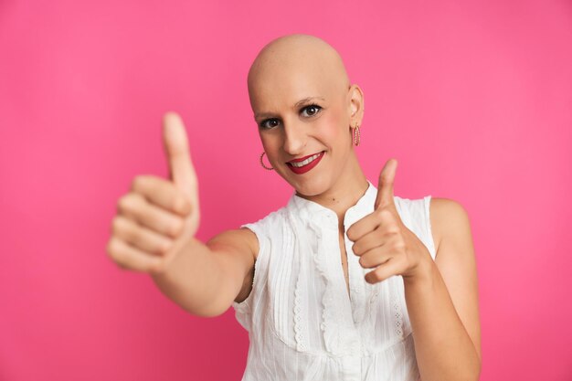 Optimistic woman suffering from cancer with shaved head and\
positive gesture on pink background