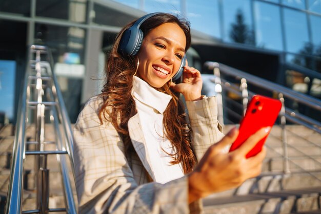 Optimistic woman wearing headphones and holding phone outdoors Communication in social networks