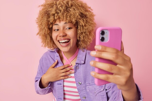 Optimistic woman with curly bushy hair takes selfie or has\
video talk poses at front camera of smartphone keeps hand on chest\
and smiles sincerely wears purple jacket isolated over pink\
background