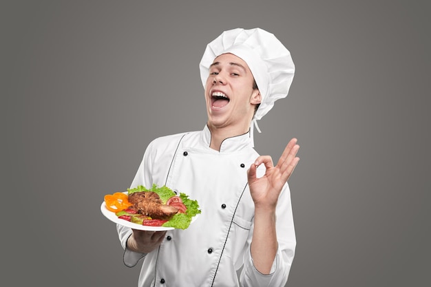 optimistic young male chef in white uniform showing delicious dish and gesturing ok against gray background