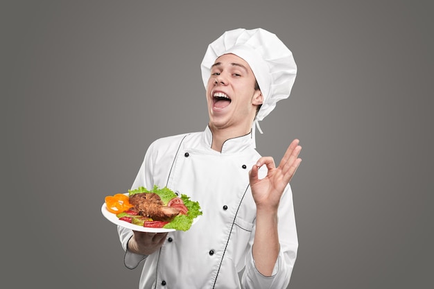 Optimistic young male chef in white uniform showing delicious dish and gesturing OK against gray background