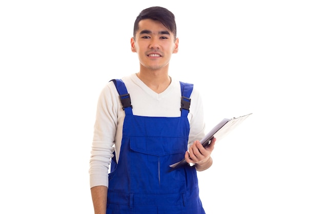 Optimistic young man with dark hair wearing in white shirt and blue overall holding white pen and black folder on white background in studio