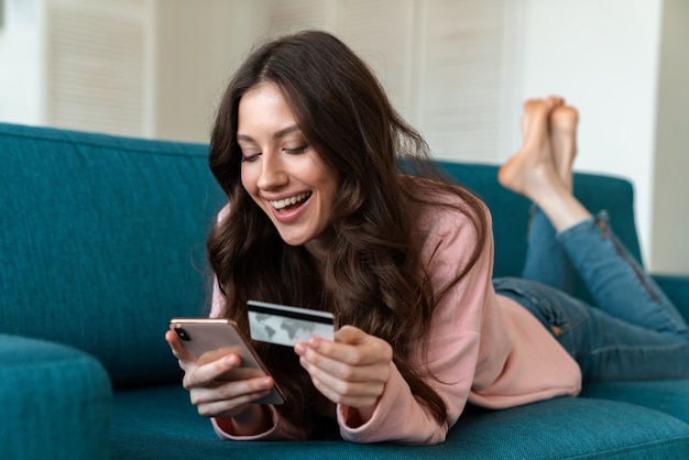 optimistic young woman indoors at home using mobile phone holding credit card on sofa.