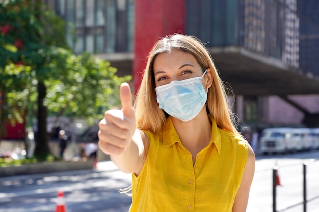 Optimistic young woman wearing protective surgical mask showing thumbs up in Sao Paulo city street Brazil