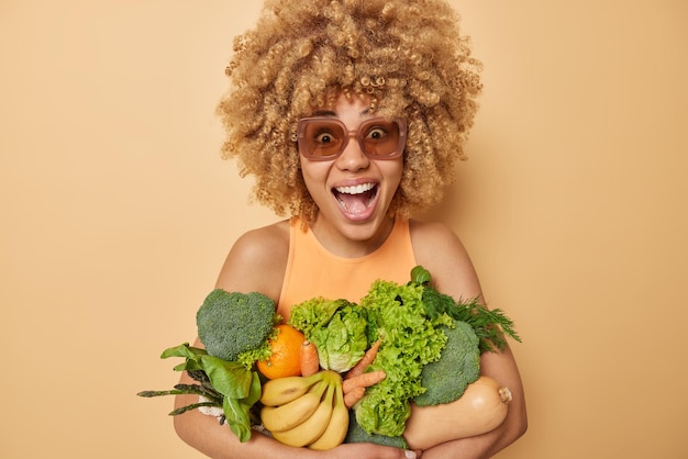 Optimistisch blij jonge vrouw met krullend haar draagt verse lente groenten rendement van markt roept luid draagt zonnebril en t-shirt geïsoleerd over beige achtergrond Groene kruidenier concept