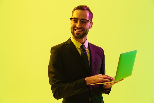 Optimistisch blij positieve gelukkige jonge zakenman poseren geïsoleerd over lichtgroene muur muur met led neonlichten met behulp van laptopcomputer.