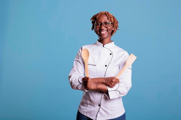 Optimistisch professioneel fornuis met houten keukengerei in studio opname. Vrouwelijke chef-kok in uniform pronkt trots op het werk en kijkt naar de camera tegen een blauwe achtergrond.