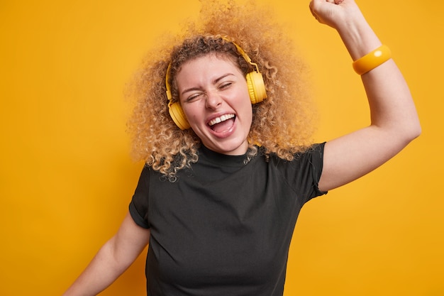 Optimistische blije europese vrouw met krullend haar heeft plezier, steekt armen op en heeft een vrolijke stemming, luistert naar favoriete muziek uit een afspeellijst die over een levendige gele muur is geïsoleerd. dolblij tienermeisje dwaast rond