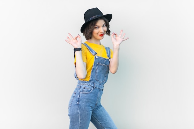 Optimistische gelukkige hipster vrouw in overall t-shirt en hoed hief haar armen op en liet een goed teken zien