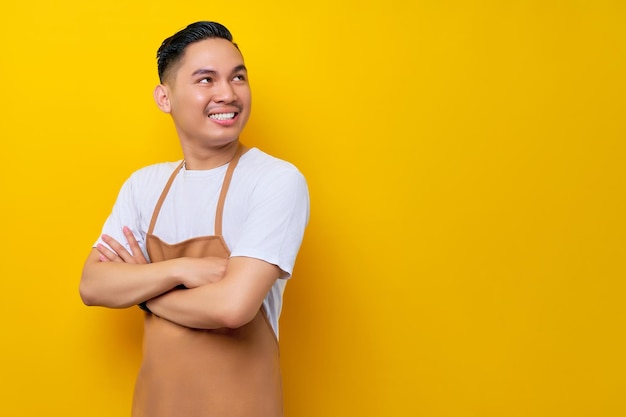 Optimistische jonge Aziatische man 20s barista-medewerker met bruine schort werkend in coffeeshop staand met gekruiste armen opzij kijkend op gele achtergrond Klein bedrijf opstarten concept