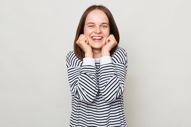 Optimistische lachende vrouw met bruin haar in een gestreept overhemd dat geïsoleerd over een grijze achtergrond staat, houdt de handen in de buurt van het gezicht terwijl ze naar de camera kijkt en gelukkig is