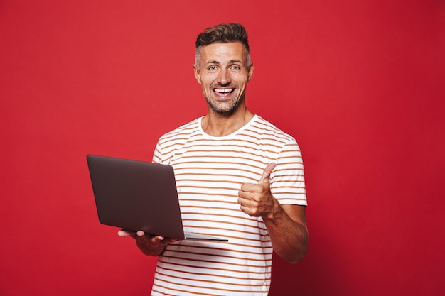 Optimistische ongeschoren man in gestreept t-shirt glimlachend en met grijze laptop geïsoleerd op rood