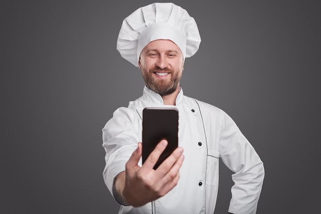 Optimistische volwassen bebaarde professionele kok in witte chef-kok uniforme selfie op mobiele telefoon tegen een grijze achtergrond