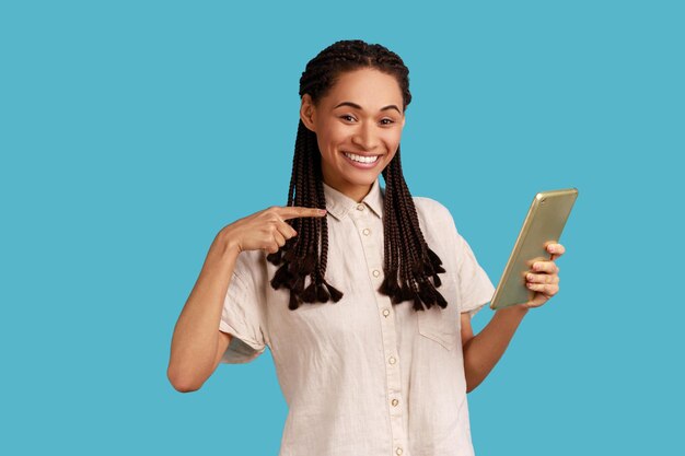 Optimistische vrouw met dreadlocks met behulp van tablet voor online werken kijken glimlachend naar camera wijzend op display dragen wit overhemd Indoor studio shot geïsoleerd op blauwe achtergrond