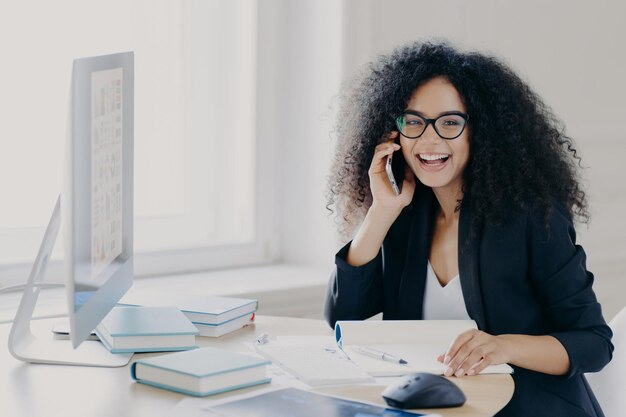 Optimistische vrouwelijke Afro-Amerikaanse ondernemer met krullend kapsel houdt mobiele telefoon vast en geniet van telefoongesprek met collega werkt op computer poseert op werkplek met notitieblok en boeken