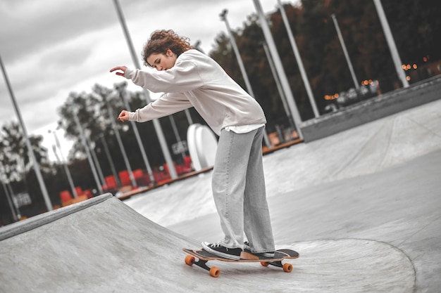 Opwaartse beweging. Meisje met golvend haar in sweatshirtjeans en sneakers met uitgestrekte armen op skateboard dat de helling op glijdt in skatepark