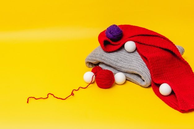 Foto su uno sfondo arancione c'è un maglione piegato grigio e una sciarpa rossa, ci sono palline rosse e blu di filo di lana, palline bianche sparse