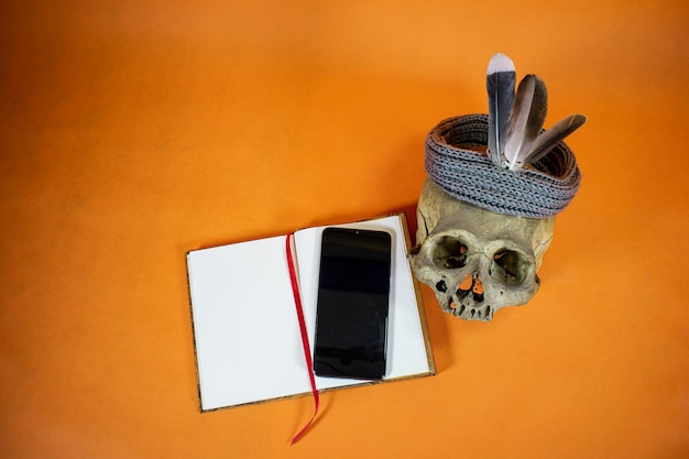 オレンジ色の背景に開いたノート、上に携帯電話、羽の装飾が施された頭蓋骨。