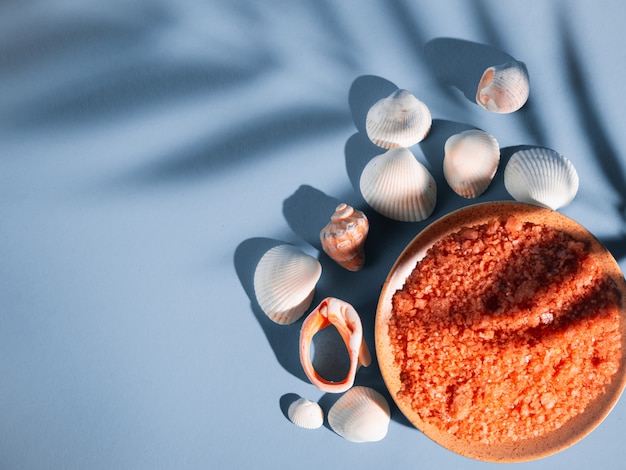 Orange bath salt in a saucer with shells on a blue background with a shadow from a tropical plant. Copyspace, flatlay. Spa, relaxed, summer