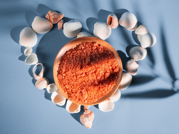 Photo orange bath salt in a saucer with shells on a blue background with a shadow from a tropical plant. copyspace, flatlay. spa, relaxed, summer