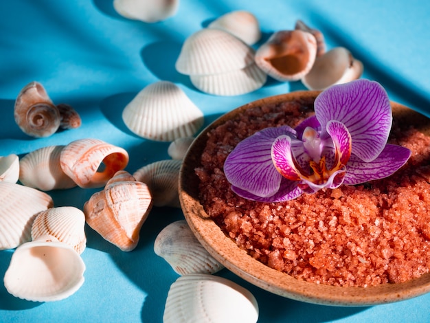 Orange bath salt in a saucer with shells and flower on a blue background with a shadow from a tropical plant. Copyspace. Spa, relaxed, summer