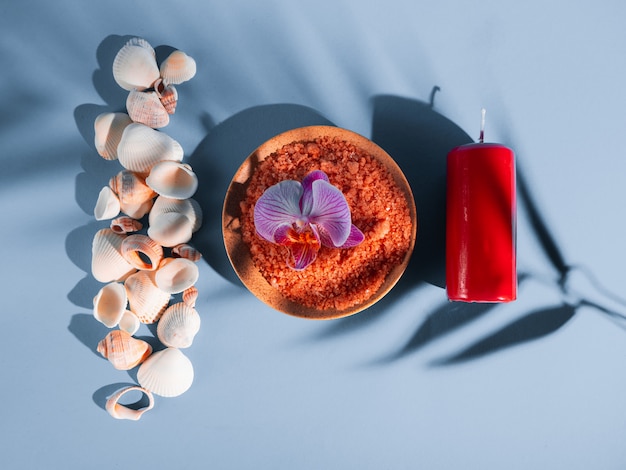 Оранжевая соль для ванн в блюдце с раковинами, красная свеча и цветок на синем фоне с тенью от тропического растения. Copyspace, flatlay. Спа, отдых, лето