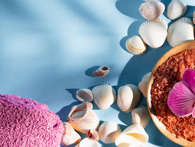 Orange bath salt in a saucer with shells, towel and flower on a blue background with a shadow from a tropical plant. Copyspace, flatlay. Spa, relaxed, summer