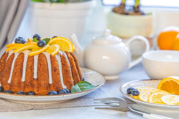 Orange bundt cake with blueberry surrounded fruits plant and cutlery on light table near window Family breakfast concept