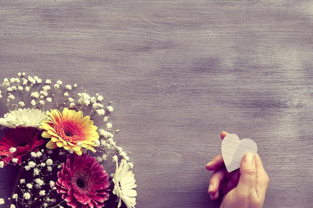 Orange and burgundy gerberas and white baby breath flowers on grey wood background with copy space place for your text Flower arrangement floral corner composition Hand hold paper heart