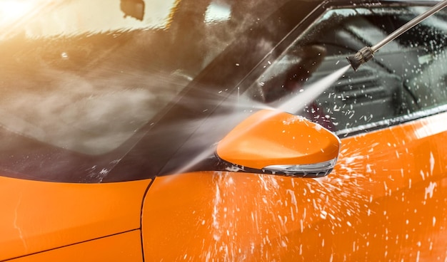 Orange car washed in self serve carwash, white shampoo and foam spraying to side mirror