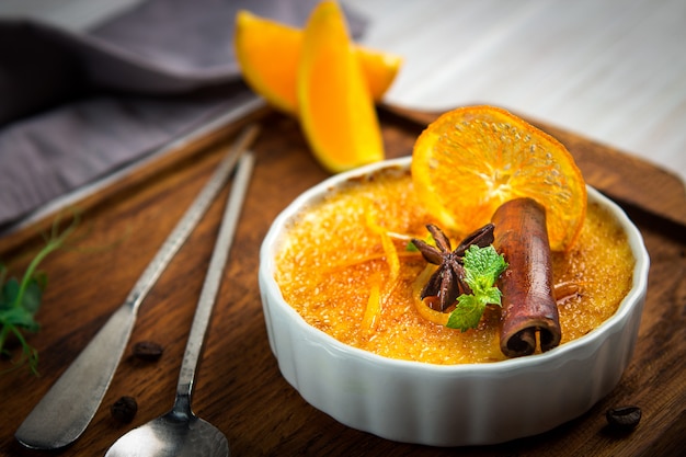 Dessert arancio della crème-brulée in ciotola bianca con cannella e menta su fondo di legno