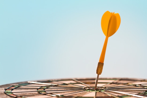 Orange dart arrow hitting bulls-eye in center target of dartboard on blue sky background with copy space